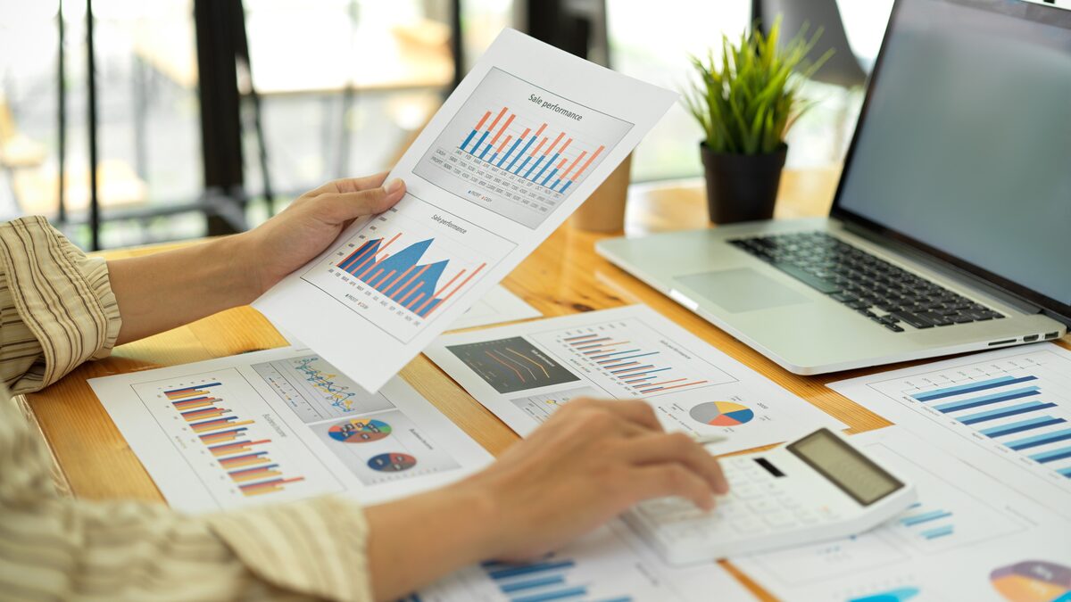 Persona revisando informes con gráficos y una calculadora junto a una laptop